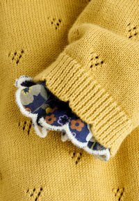 Žltý pletený sveter s textúrovaným vzorom a volánikovým kvetinovým lemom na manžetách. Materiál je mäkký s hrubým pletením.