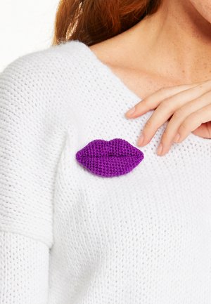 Ręka w białym, dzianym swetrze z przypiętą obok obojczyka fioletową broszką w kształcie ust wykonanych na szydełku.