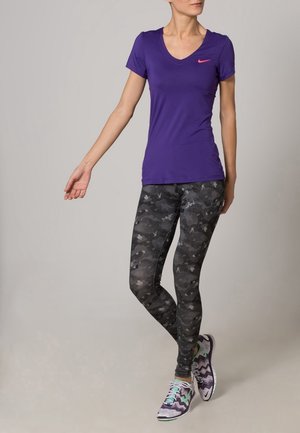 Tee-shirt de sport violet à manches courtes avec col en V, associé à un legging camouflage noir et des chaussures de course multicolores avec un motif ondulé.