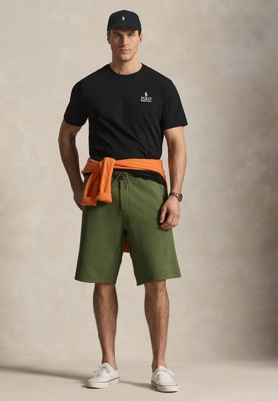 Vīrietis stāv, valkājot melnu Polo T-kreklu, zaļas šortus, baltus sporta apavus, melnu cepuri un oranžu džemperi, kas piesiets ap vidukli, neitrāla fona priekšā.