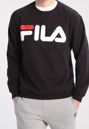 Person iført sort FILA-sweatshirt med stort logo og lysegrå joggingbukser med lille logo, hænder i lommerne, stående foran en ensfarvet baggrund.