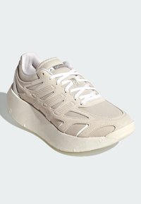 Beige idrottssko med ovandel i mesh och mocka, tjock vit mellansula, texturerade detaljer och snörningsdesign. Har logotyp på tungan.