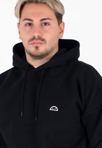 Czarna bluza z kapturem o strukturze w jodełkę, z małym haftowanym logo w białym i niebieskim kolorze na piersi. Kaptur z regulowanym sznureczkiem.