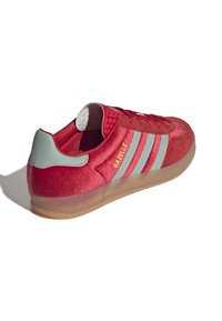 Baskets en suede rouge avec des accents verts, trois bandes blanches, et une semelle en caoutchouc translucide. Comprend un col rembourré et une languette texturée.