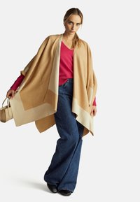 Donna in piedi che indossa un poncho beige e crema sopra un maglione rosso, jeans blu a gamba larga, scarpe nere, tenendo una piccola borsa beige.