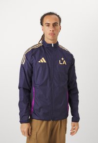 Cazadora atlética color navy con acentos y rayas doradas, que cuenta con cierre completo y logotipo de LA en el pecho, combinada con pantalones caqui.