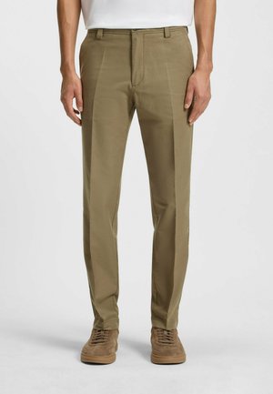 Pantalons khaki avec une texture lisse, design à jambes droites et devant plat. Portés avec des baskets en daim marron. Style minimaliste.
