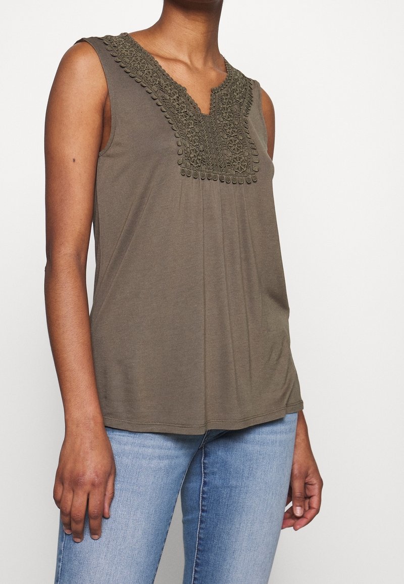 Haut sans manches vert olive avec un motif floral crocheté complexe au col en V, porté avec un jean bleu clair sur un modèle à la carnation moyenne.