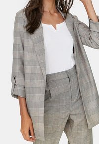 Grijze geruite blazer met opgerolde mouwen, gecombineerd met een witte top. De blazer heeft een opstaande kraag en een relaxte pasvorm, benadrukt lijnen en textuur.