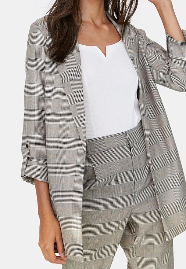 Grijze geruite blazer met opgerolde mouwen, gecombineerd met een witte top. De blazer heeft een opstaande kraag en een relaxte pasvorm, benadrukt lijnen en textuur.