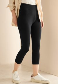 Zwarte capri legging gemaakt van rekbaar materiaal, met een hoge tailleband en een zachte textuur. Gedragen met witte sportschoenen.