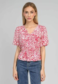 Blouse à motifs rouges et blancs avec un décolleté en V et des manches courtes, avec une taille smockée et un ourlet légèrement évasé. Portée avec un jean bleu.