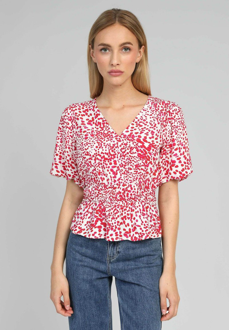 Blouse à motifs rouges et blancs avec un décolleté en V et des manches courtes, avec une taille smockée et un ourlet légèrement évasé. Portée avec un jean bleu.