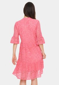 Rochie roz din dantelă, cu un design cu mâneci evazate, tiv ondulat și modele florale, evidențiind o textură semi-transparentă.