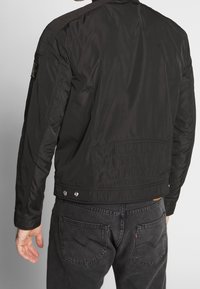 Svart bomberjacka med slät yta, elastiska ärmslut och dragkedja fram. Har sidofickor och ett logopatch på vänster axel.