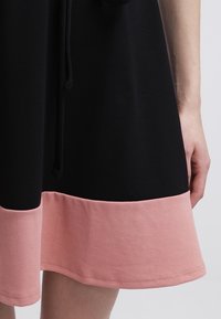 Robe noire avec un ourlet rose. Présente une texture douce, un cordon de serrage à la taille et une coupe décontractée. Les blocs de couleur sont distincts et bien définis.