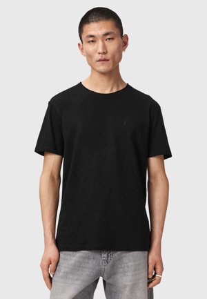 Jeune homme aux cheveux courts portant un t-shirt noir à col rond et un jean gris clair, debout devant un fond uni gris clair.