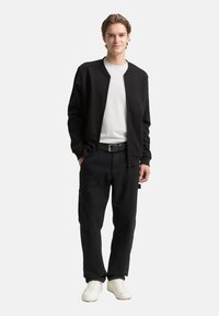 Schwarze Bomberjacke mit Reißverschluss über einem weißen T-Shirt, kombiniert mit schwarzen Hosen und weißen Sneakers. Merkmale sind gerippte Bündchen und Bund.