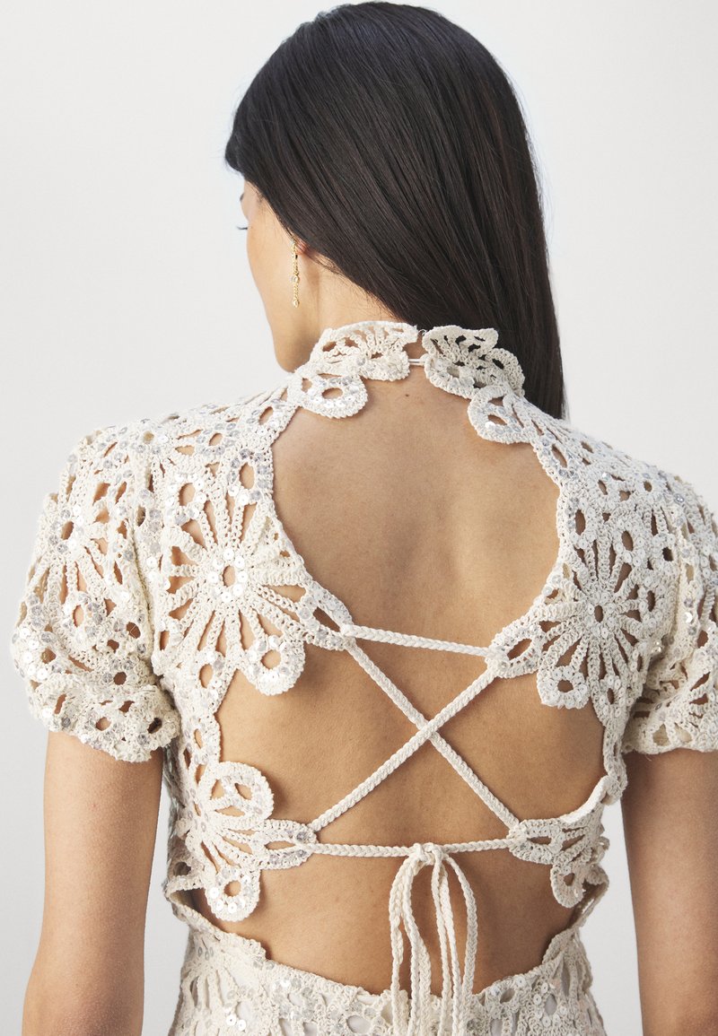 Gehäkeltes Oberteil in hellem Beige mit floralen Mustern, kurzen Puffärmeln, einem offenen Rücken mit gekreuzten Bändern und strukturierten Details.