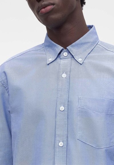 Vue partielle d'une personne portant une chemise Oxford bleu clair à boutons avec un col boutonné et une poche poitrine.