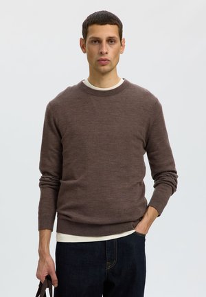 Homme portant un pull marron et un jean foncé, debout avec une main dans la poche et tenant un sac dans l’autre, sur un fond clair uni.