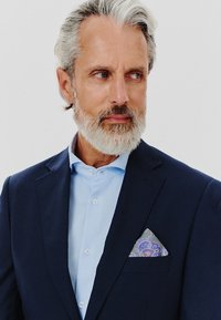 Granatowa marynarka z jasnoniebieską koszulą, z prostokątną kieszonką w kolorowy wzór paisley. Krótkie szare włosy i broda.
