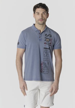 Hombre con polo azul con texto gráfico y pantalones cortos blancos, de pie contra un fondo claro liso.