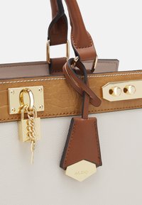 Sac à main avec un corps crème texturé, des accents marron, une quincaillerie dorée et une étiquette décorative avec une chaîne et un détail de cadenas.