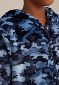 Zila kamuflāžas raksta polāra materiāla kapuce ar rāvējslēdzēju. Iezīmes: mīksta tekstūra, oderēta kapuce un tumši zils rāvējslēdzējs ar reljefu akcentu.