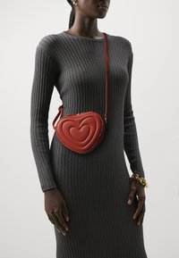 Red leather heart-shaped crossbody bag with quilted design and thin strap, worn over a gray ribbed long-sleeve dress. Gold chain bracelet visible.