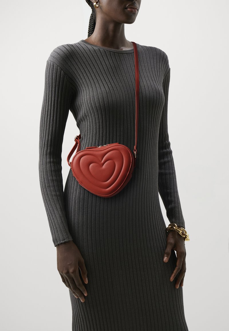Red leather heart-shaped crossbody bag with quilted design and thin strap, worn over a gray ribbed long-sleeve dress. Gold chain bracelet visible.