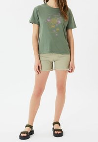 Camiseta de algodón verde con estampado gráfico floral en rosa y dorado. Combinada con pantalones cortos deshilachados de color verde claro y sandalias negras gruesas.