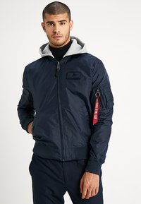 Chaqueta bomber azul marino con capucha gris, cierre de cremallera, puños de canalé y un parche en el pecho. Presenta un bolsillo lateral con acento de hardware.