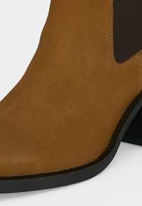 Bottine en daim marron avec un panneau latéral élastique noir et un talon bloc bas noir sur un fond clair.