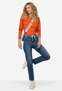 Oranssi pitkähihainen paita, vaaleansiniset farkut naruilla ja off-white-plattformitossut. Pieni vaalea laukku on kuljetettu poikittain.