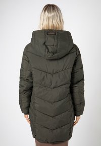 Person mit blonden Haaren, die einen langen, dunklen grünen gepolsterten Mantel mit Kapuze trägt, von hinten vor einem schlichten Hintergrund betrachtet.