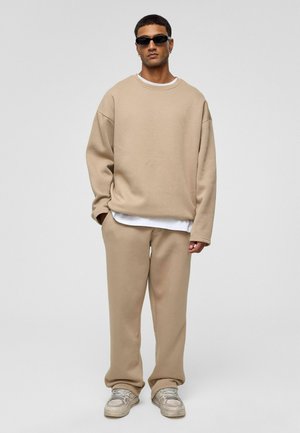 Beige oversized sweatshirt met lange mouwen, gedragen over een wit shirt, gecombineerd met bijpassende broek en grijze sneakers. Zwarte zonnebril gedragen.