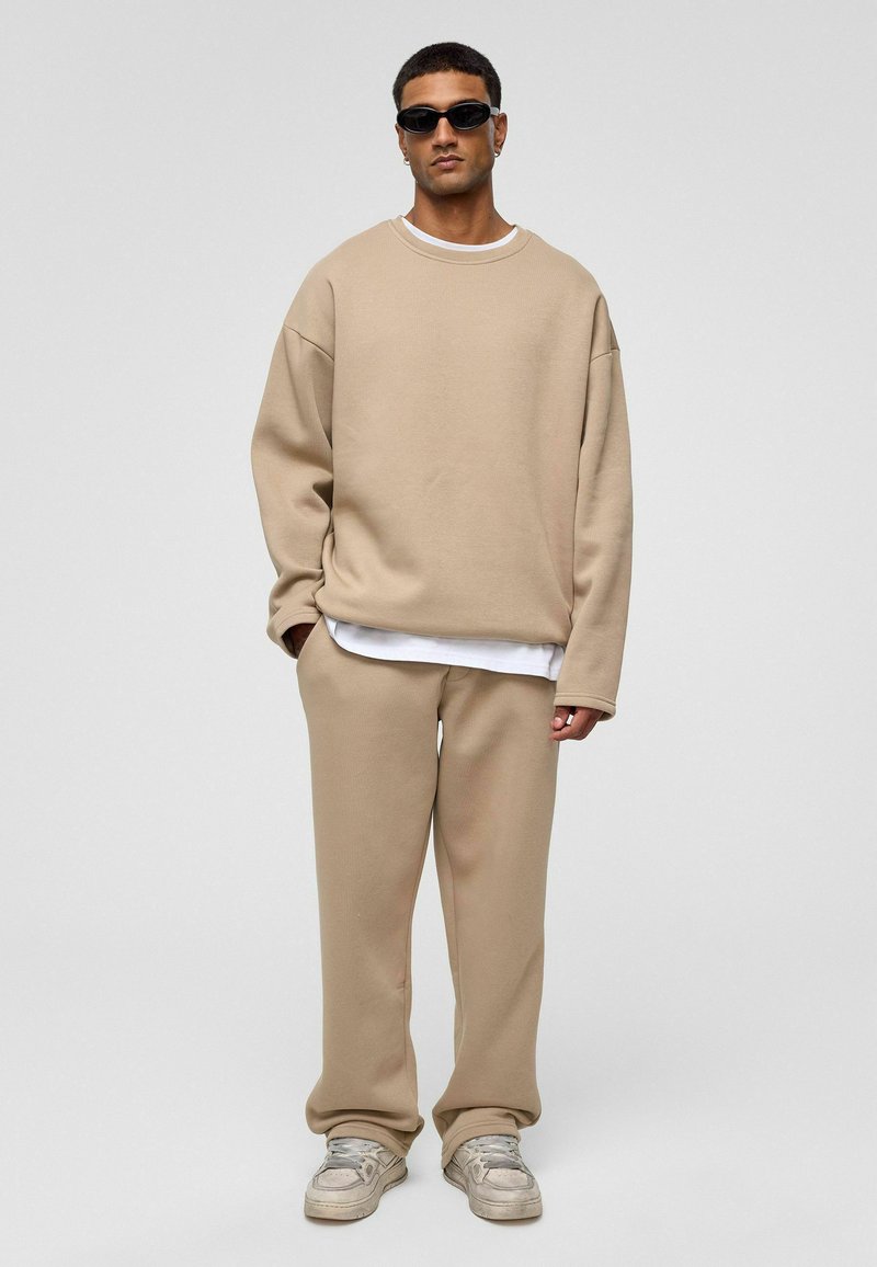 Beige oversized sweatshirt met lange mouwen, gedragen over een wit shirt, gecombineerd met bijpassende broek en grijze sneakers. Zwarte zonnebril gedragen.