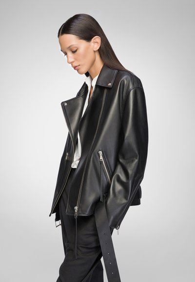 Schwarze Lederjacke mit strukturiertem Design, auffälligen silbernen Reißverschlüssen, Schulterklappen und einem leicht übergroßen Schnitt. Glatte Textur und taillierter Schnitt.
