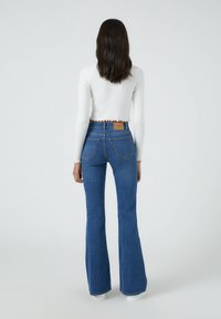 Jean flare bleu taille haute, avec deux poches arrière et une étiquette en cuir marron. Associé à un haut blanc court à manches longues.