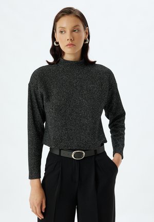 Top negro brillante de mangas largas con cuello alto, usado con un cinturón negro que tiene una hebilla redonda de plata. Tejido suave y texturizado.