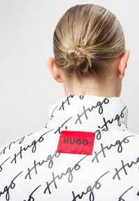 Vit puffjacka med svart "Hugo"-tryck, med en röd logotyp-patch på baksidan. Slät textur och hög krage.