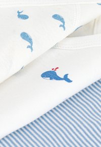 Kottongarn med blåhvalsmønster på hvit bakgrunn, med en tydelig blåhval med røde detaljer på ett plagg. Stripete stoff nedenfor.