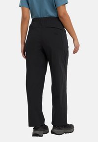 Schwarze, elastische Hose mit Taschen, gerade geschnitten und aus leichtem Stoff, kombiniert mit grauen Wanderschuhen, von hinten betrachtet.