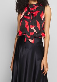 Blouse noire sans manches avec motifs de feuilles rouges et un lien à l'avant, associée à une jupe en satin noir brillant.