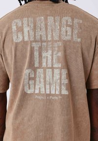 Camiseta marrón de manga corta con gran texto blanco texturizado en la parte de atrás que dice "CAMBIA EL JUEGO" y "Project x Paris™."
