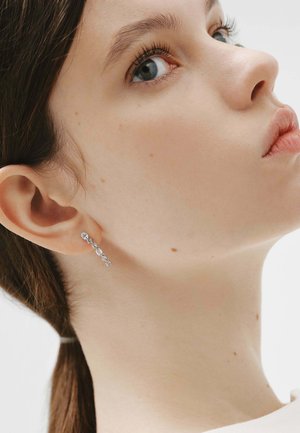 Mujer joven de piel clara y ojos azules, lleva un pendiente plateado colgante con pequeñas gemas, cabeza ligeramente elevada y girada hacia un lado.