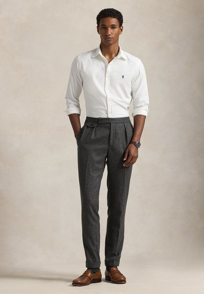 Joven de pie con las manos en los bolsillos, vistiendo una camisa blanca de botones, pantalones grises plisados, mocasines marrones y un reloj de pulsera.