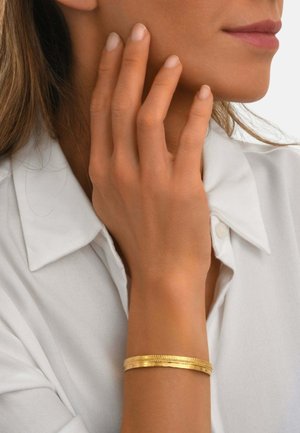 Femme portant une chemise blanche, touchant son visage de la main gauche, montrant un bracelet fin en or texturé à son poignet.