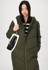 Abrigo acolchado verde con cremallera, capucha y diseño acolchado. Parte superior a rayas en blanco y negro, llevando un bolso con una correa blanca esponjosa y un cuerpo negro brillante.